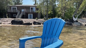 Una silla Adirondack azul en agua cristalina y poco profunda, con una casa y una hamaca al fondo.