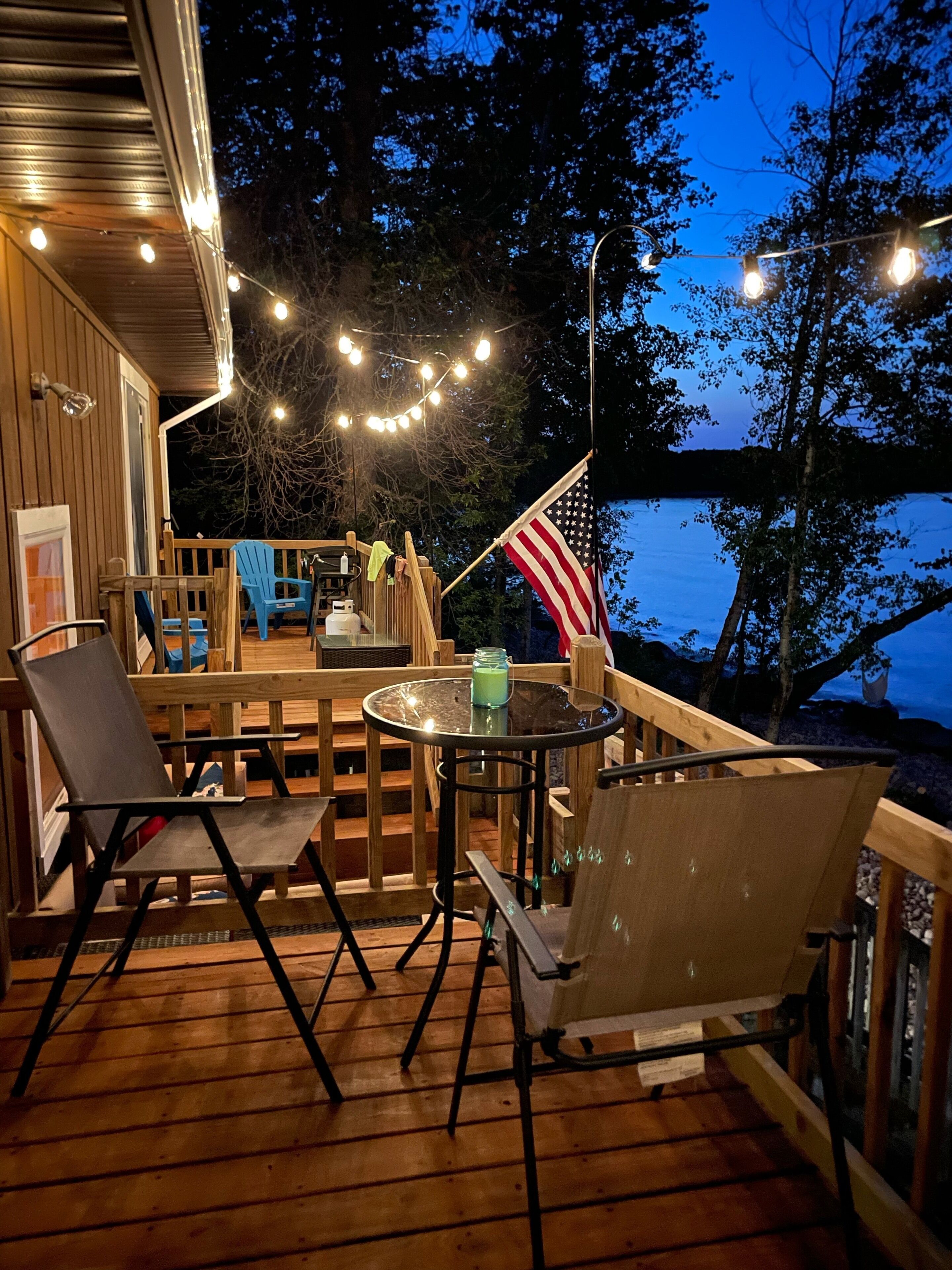 Eine Terrasse im Freien mit Holzboden, Metallstühlen, einem runden Tisch mit Glasplatte, Lichterketten und einer US-amerikanischen Flagge.