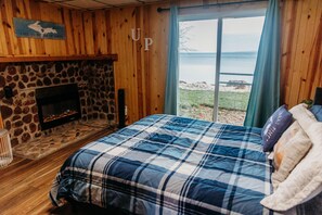Ein Schlafzimmer mit Kamin aus Stein, einem Bett mit blau-weiß karierter Bettdecke und Blick auf den See durch ein großes Fenster.