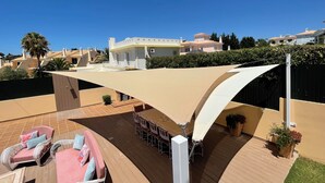Uma área de pátio com móveis de exterior, um guarda-sol com sombra e vista para um prédio residencial ao fundo.
