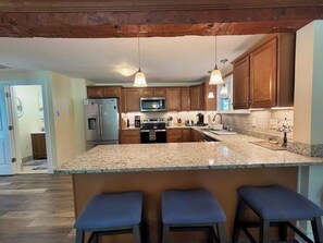 A kitchen with wooden cabinets, a granite countertop, a stainless steel refrigerator, a microwave, and a stove. There are three bar stools and pendant lights above the counter.