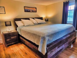 A bedroom with a wooden bed frame, a headboard, and a large window with curtains. - Beau Rivage Waterfront, kayak, SUP, great fishing, swimming, fire pit, updated (Portlandville)