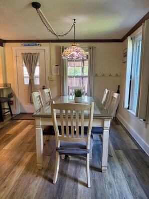 A dining area with a glass table, wooden chairs, a hanging light fixture, and a window with curtains. - Beau Rivage Waterfront, kayak, SUP, great fishing, swimming, fire pit, updated (Portlandville)