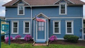 A two-story blue house with a front porch, a small American flag, and pink flowering bushes. - Beau Rivage Waterfront, kayak, SUP, great fishing, swimming, fire pit, updated (Portlandville)