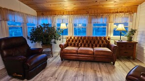 A cozy living room with a leather sofa, a leather armchair, and a wooden floor. There are two windows with curtains, a potted plant, and a lamp on a side table. - Beau Rivage Waterfront, kayak, SUP, great fishing, swimming, fire pit, updated (Portlandville)