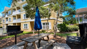 Una terraza de madera con mesas de picnic y una parrilla, rodeada por un edificio amarillo con balcones.