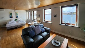 Een moderne kamer met een houten plafond, een zwarte leren bank, een bed en grote ramen met uitzicht op een waterpartij.