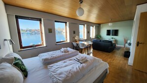 Hotelkamer met een groot bed, een bank, een eettafel en uitzicht op een waterlichaam door grote ramen.