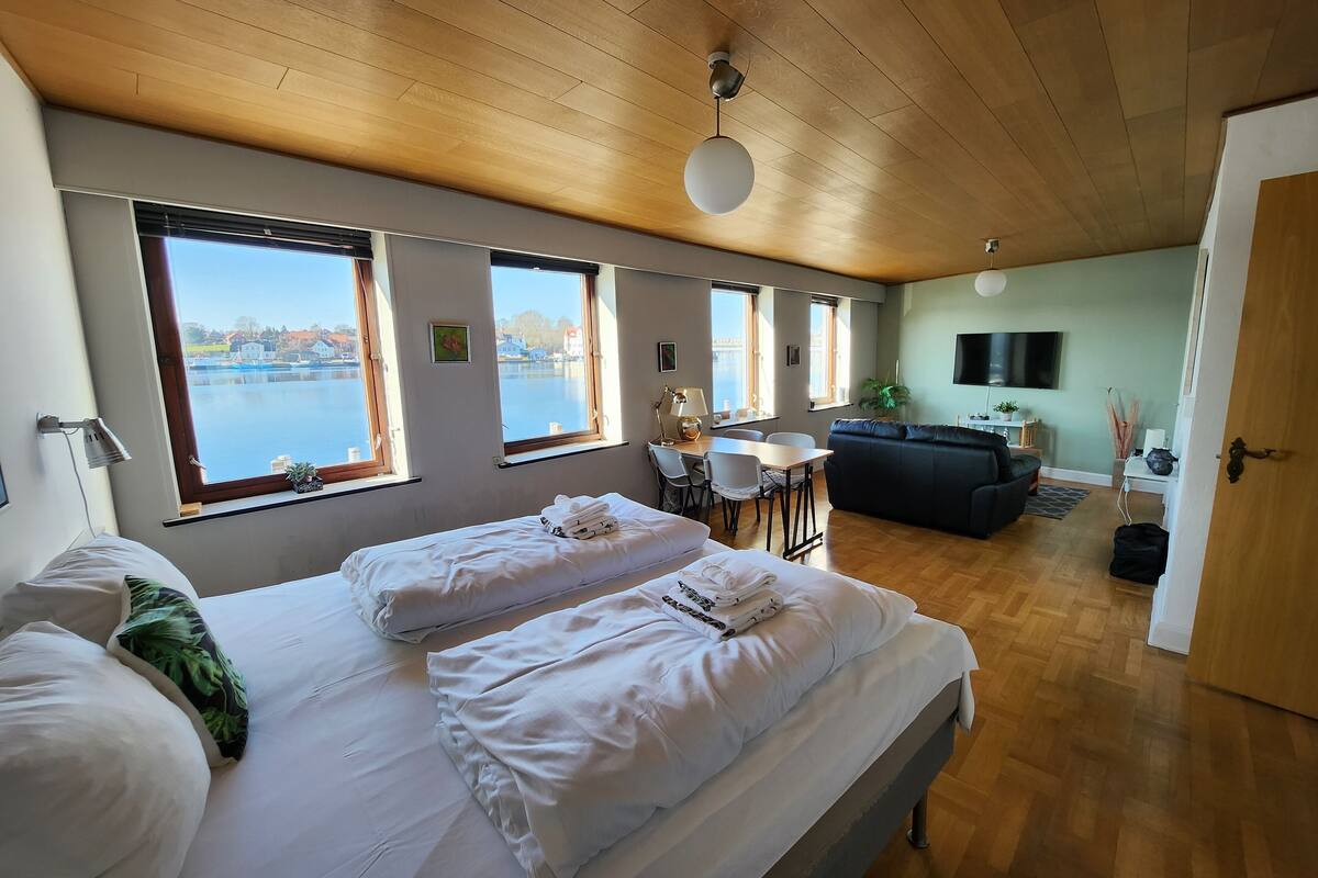 Habitació d'hotel amb un llit gran, un sofà, una taula de menjador i vista a un cos d'aigua a través de grans finestres.
