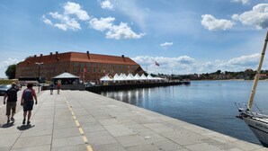 Een waterkantpromenade met een groot bakstenen gebouw, een rij witte tenten en een rustig waterlichaam.