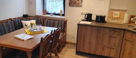 A dining area with a wooden table and chairs, a window with a view of greenery, and a kitchen with wooden cabinets and a sink.