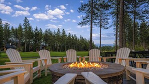 Un hogar circular rodeado de sillas Adirondack de madera, situado en un  área verde al aire libre con árboles y cielo despejado.
