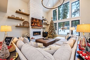 A cozy living room with a fireplace, a Christmas tree, and a large window with a view of trees.