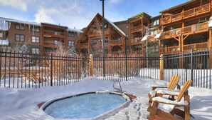 Een besneeuwde buitenruimte met een hottub, houten Adirondack-stoelen en een gebouw met balkons.