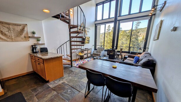 Une cuisine chaleureuse avec une table en bois, des chaises, un escalier en colimaçon et de grandes fenêtres donnant sur l’extérieur.