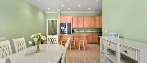 Un comedor con una mesa y sillas blancas, un jarrón de flores y una cocina de fondo con armarios de madera y un refrigerador.