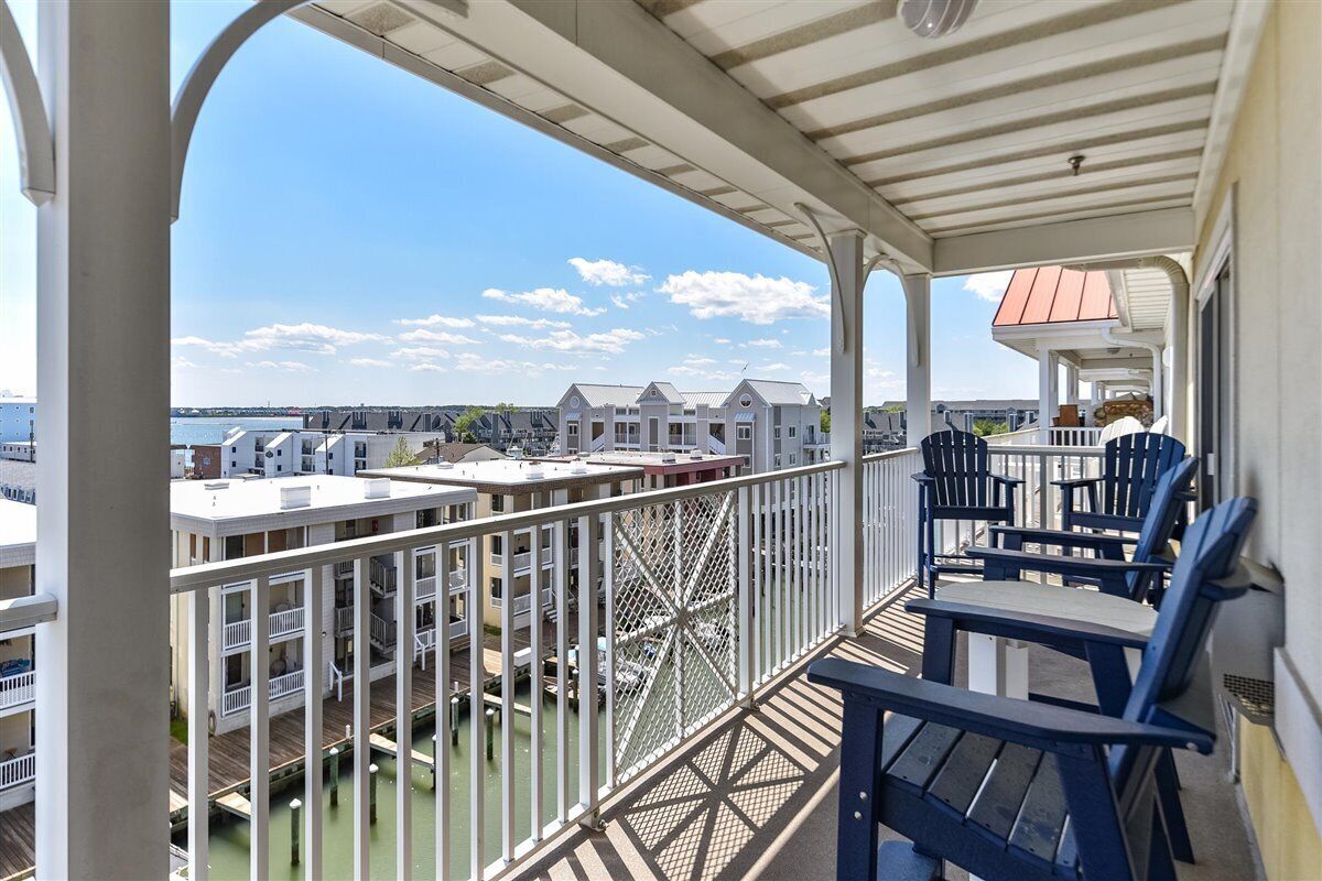 A covered balcony with a view of a marina and buildings, featuring a railing and several Adirondack chairs.