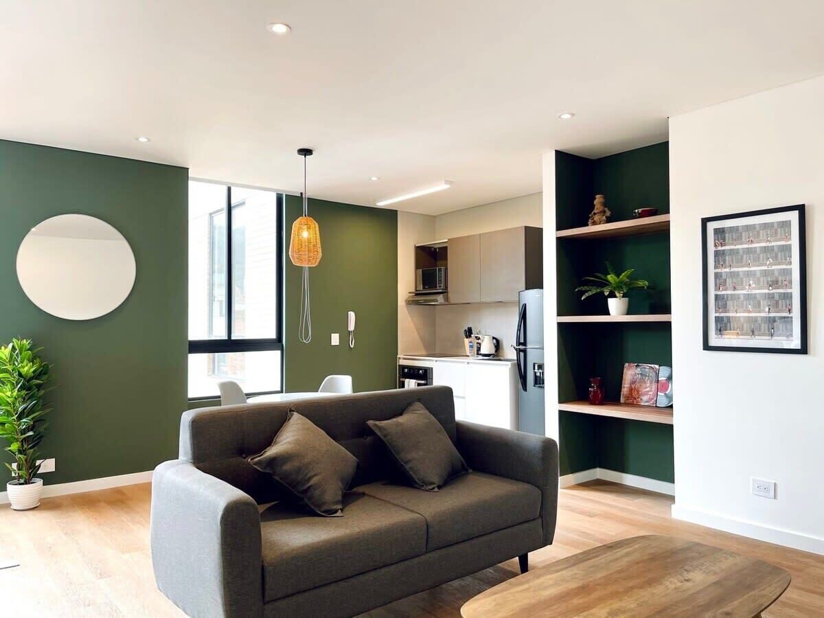 A modern living room with a grey sofa, a hanging light fixture, and a kitchen in the background.