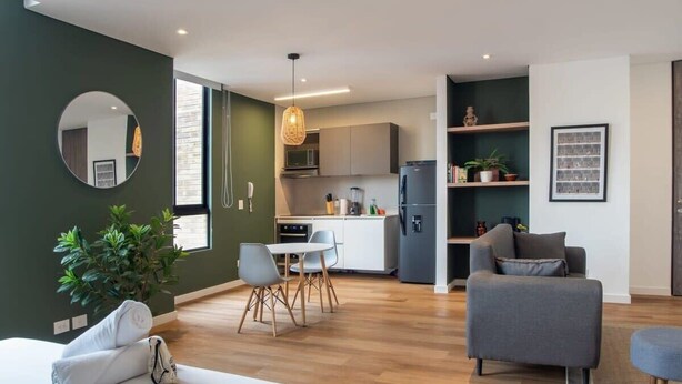 A modern living room with a sofa, a dining table with chairs, a kitchen area with cabinets and appliances, and a large mirror on the wall.