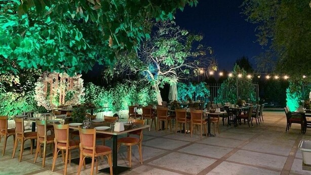 An outdoor dining area with tables and chairs, surrounded by greenery and illuminated by string lights. - Rami Hotel (Falougha-Khalouat)