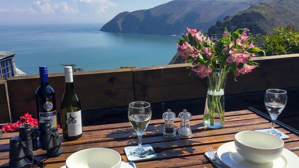 Una mesa de madera dispuesta con botellas de vino, copas, un florero con flores y cuencos, con vistas panorámicas del mar y las colinas.