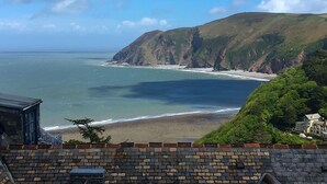 Vista costera con un muro de piedra en primer plano, una playa y un acantilado con vegetación.