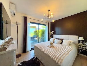 A bedroom with a large bed, a chandelier, a nightstand, and a view through a glass door to the outdoors. - Magnificent villa with heated pool air conditioning (Viviers-du-Lac)