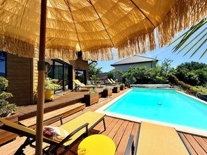 A swimming pool with wooden deck and lounge chairs, surrounded by greenery and a thatched umbrella.