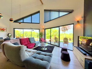 A spacious living room with a grey sofa, red chairs, a coffee table, and a TV. Large windows offer a view of greenery outside. - Magnificent villa with heated pool air conditioning (Viviers-du-Lac)