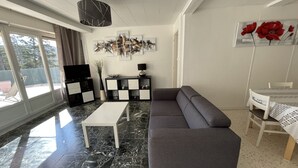 Ein modernes Wohnzimmer mit einem grauen Sofa, einem weißen Couchtisch und einer schwarzen TV-Konsole. Es gibt einen Essbereich mit Tisch und Stühlen, und ein großes Fenster bietet einen Blick ins Freie.