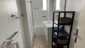 Ein schmaler Flur führt zu einem Badezimmer mit einem weißen Heizkörper, einem kleinen Fenster und einem Spiegel über einem Regal mit Körben.