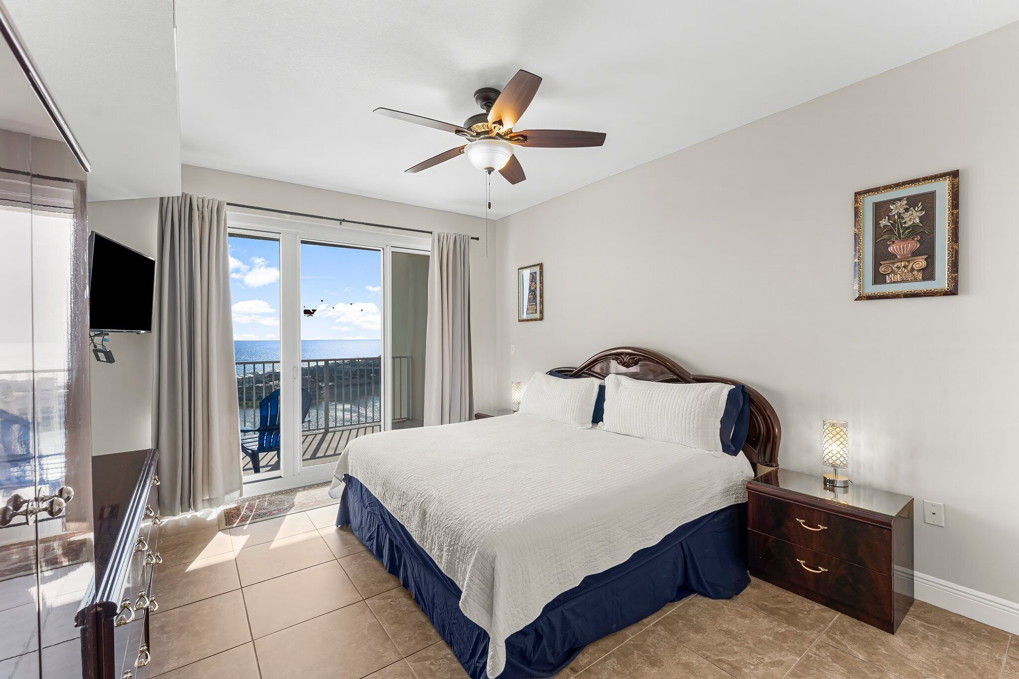 A hotel room with a large bed, a ceiling fan, a nightstand with a lamp, a TV, and a balcony with a view of the ocean.