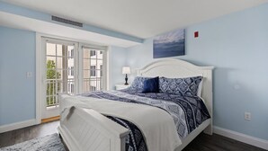 Un dormitorio con cama blanca, ropa de cama estampada, una ventana con vistas y un cuadro en la pared.