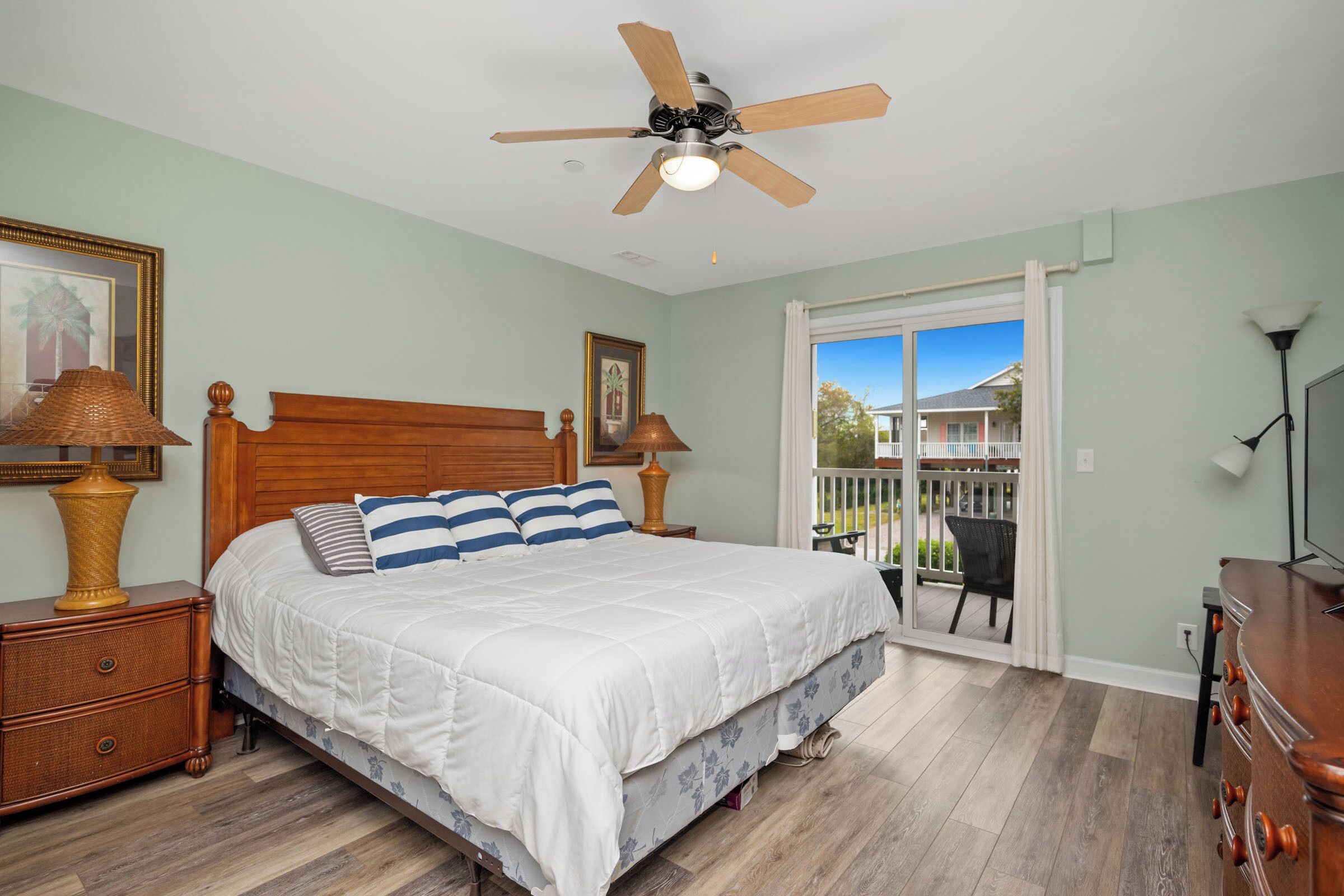 A bedroom with a bed, wooden headboard, ceiling fan, and a view of the outside.