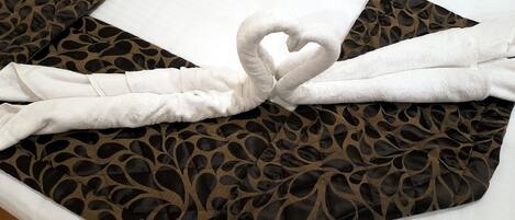 Uma cama com colcha estampada, arranjo de toalhas em formato de coração e travesseiro branco.