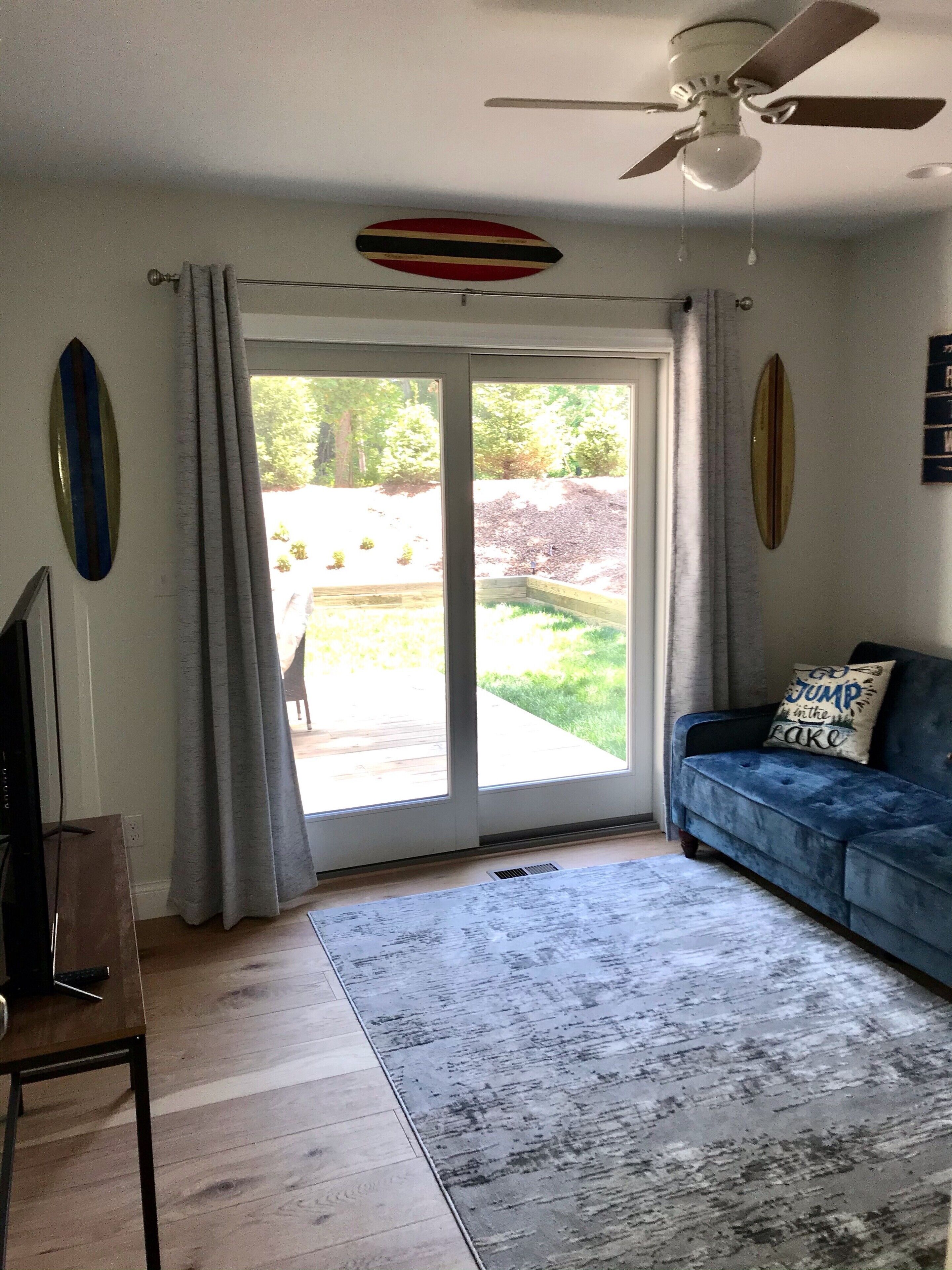 Un salon avec un canapé bleu, un ventilateur de plafond, un téléviseur et une porte-fenêtre coulissante donnant sur un jardin.
