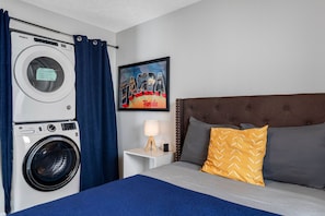 Habitación de hotel con cama, mesita de noche con lámpara, lavadora y un cuadro en la pared.