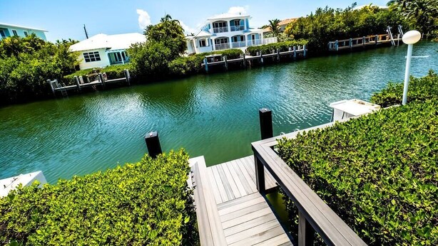 A waterfront property with a dock, a white house with a blue roof, and lush greenery. - *Newly Renovated Single Family Home w/Private Pool (Key Colony Beach)