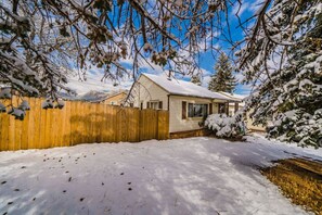 A snow-covered backyard with a wooden fence, a house with a gabled roof, and snow-laden trees.