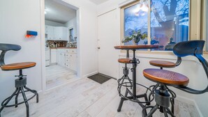 Un comedor con dos taburetes y una mesa, una cocina al fondo y una ventana con vista a árboles.