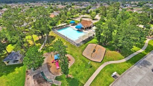 Vista aérea de una zona residencial con una piscina grande, parque infantil y área de picnic cubierta, todo rodeado de árboles y casas.