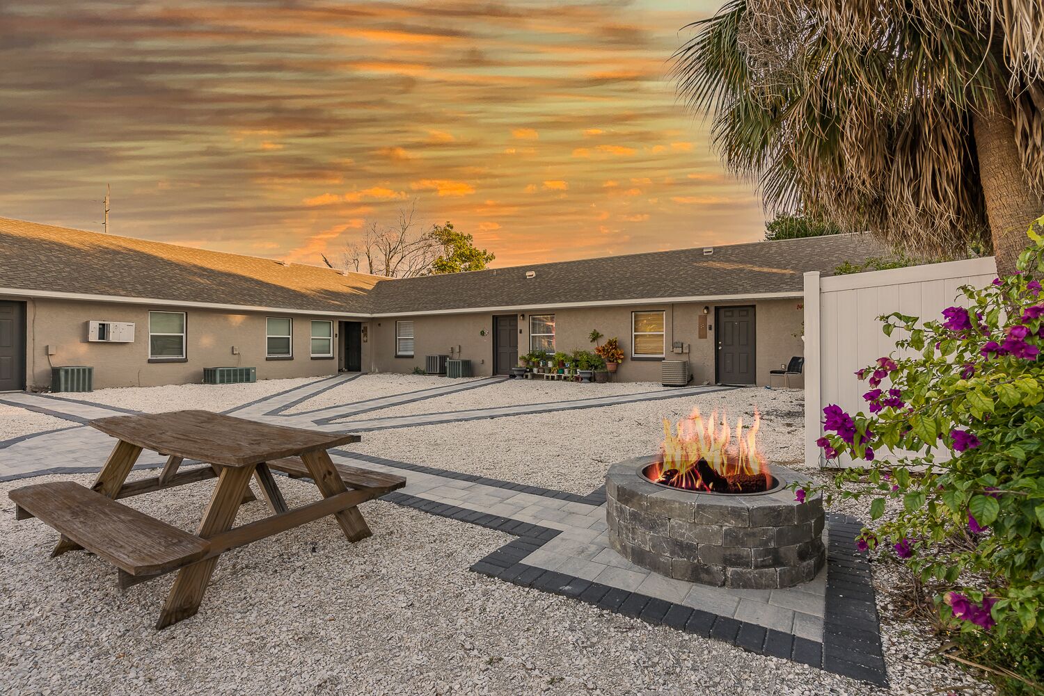 A courtyard with a fire pit and picnic table, surrounded by buildings with a gravel path.