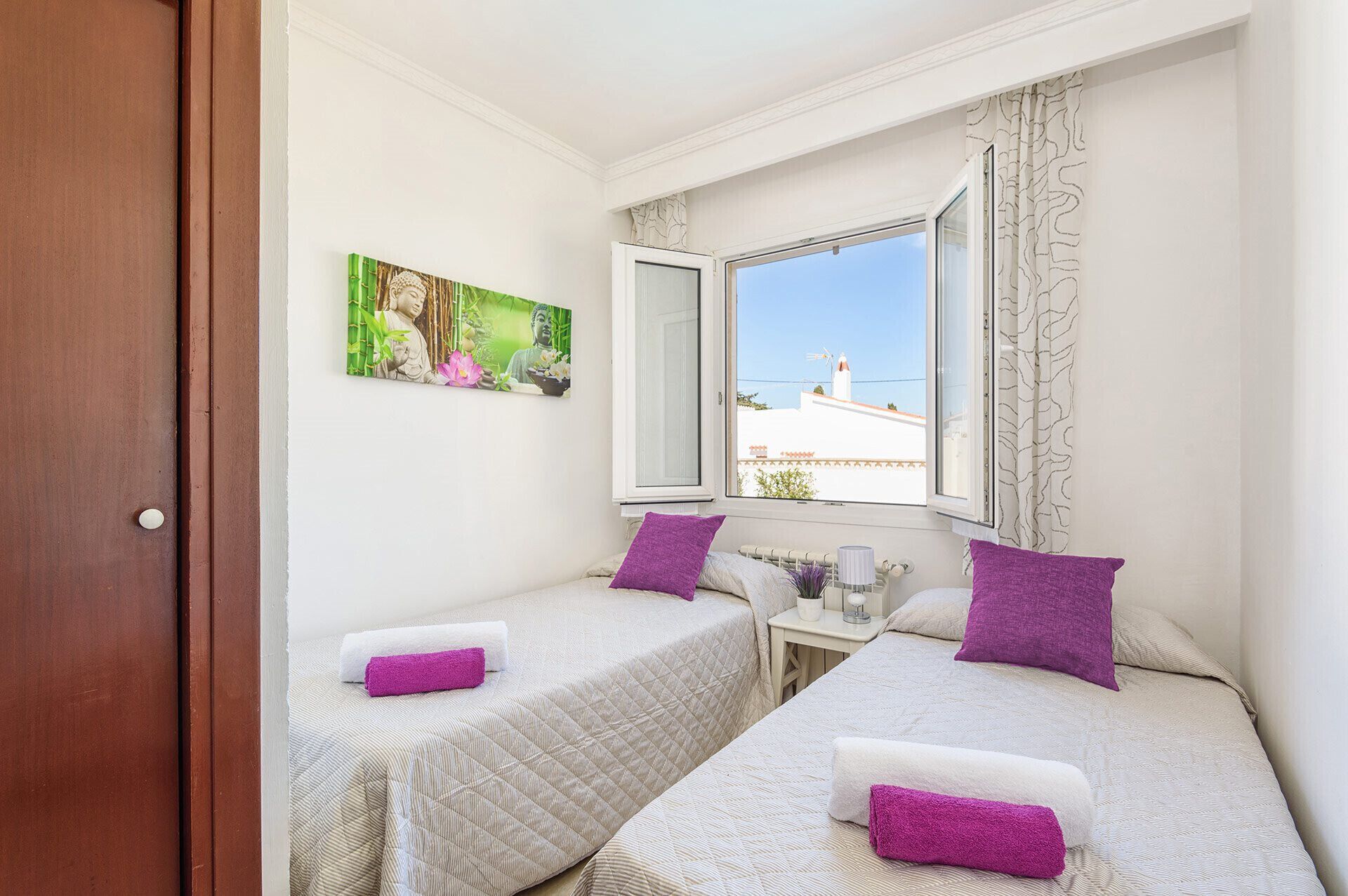 A bedroom with two beds, a window with a view, a painting on the wall, and a door.