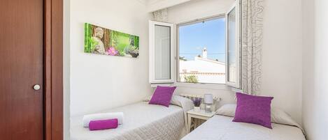 A bedroom with two beds, a window with a view, a painting on the wall, and a door.