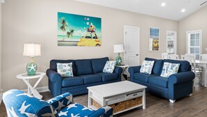 A living room with two blue sofas, a white coffee table, and a painting of a car on a beach.
