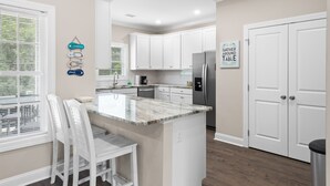 A modern kitchen with white cabinets, a granite countertop, and a stainless steel refrigerator. There is a dining area with white chairs and a small table.