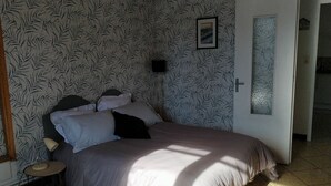 A bedroom with a bed, a bedside table, a lamp, a door, and a wall with a leaf pattern. - En Ville Avec un air de Campagne (Arles)