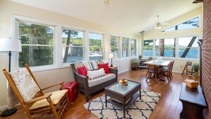 A spacious living room with a large window overlooking a body of water, a dining area with a table and chairs, and a cozy seating area with a sofa and a rocking chair. - Lake House with gorgeous view, private beach, dock space, fireplace, hammock (Bolton Landing)