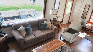 A living room with a brown sofa, a wooden coffee table, and a white armchair. - Lake House with gorgeous view, private beach, dock space, fireplace, hammock (Bolton Landing)