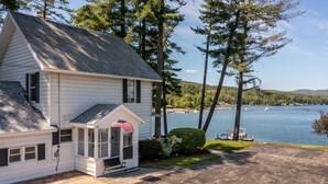 A white house with a porch, an American flag, and a view of a lake with boats. - Lake House with gorgeous view, private beach, dock space, fireplace, hammock (Bolton Landing)